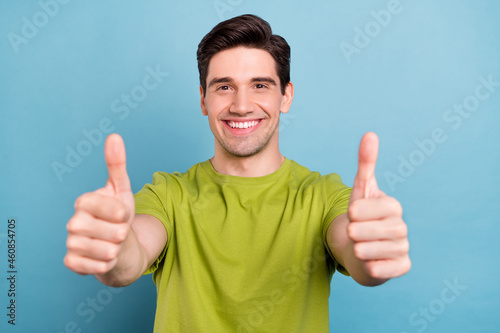 Photo of adorable sweet young man wear green t-shirt smiling showing you thumbs up isolated blue color background © deagreez