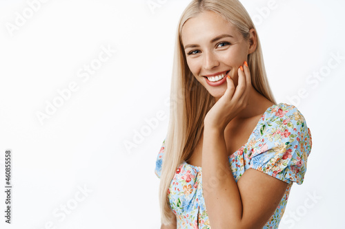 Skin care and women beauty. Attractive blond young woman touching her face expression, gazing romantic at camera and smiling, standing over white background