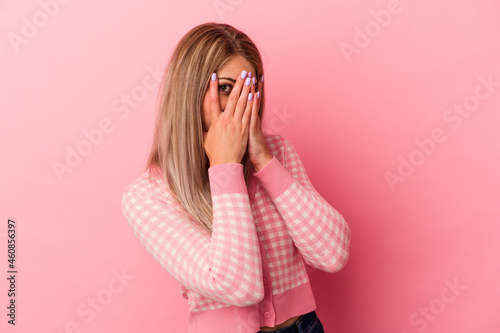 Young russian woman isolated on pink background blink through fingers frightened and nervous.