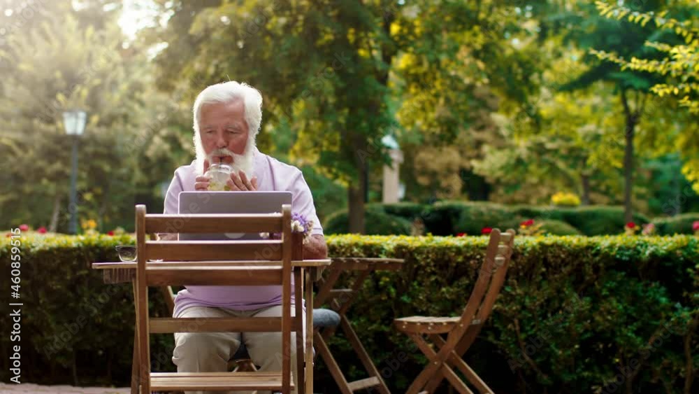 Charismatic old man at the cafe in the middle of the park Woking on his laptop typing something concentrated Charismatic old man at the cafe in the middle of the park Woking on his laptop typing something concentrated