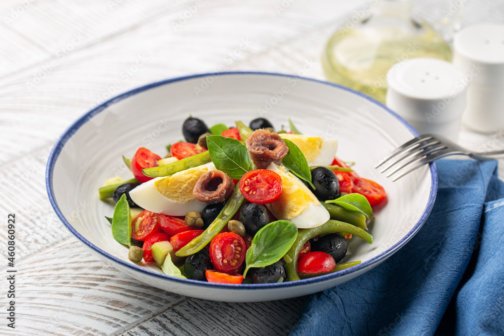 Close-up of mediterranean salad with anchovy, green beans, tomatoes, eggs, black olives, capers on white plate and white wooden background. Healthy food, no carb diet concept.
