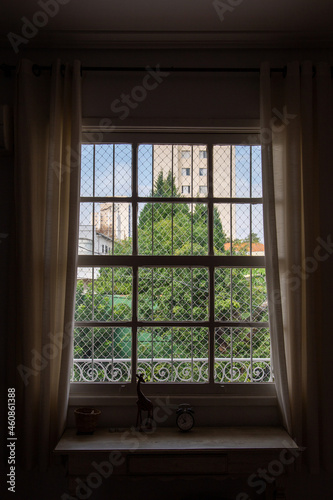 window with grid with home garden background and white curtain. With sideboard furniture and an old clock