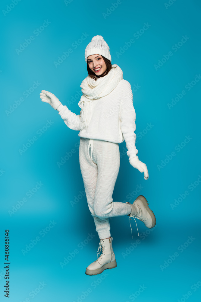 Smiling woman in white sweater, scarf and hat looking at camera on blue background Smiling woman in white sweater, scarf and hat looking at camera on blue background