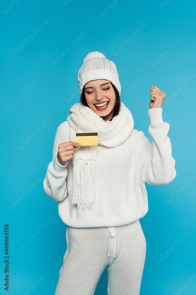 Excited woman in white knitted clothes showing yes gesture and holding credit card isolated on blue Excited woman in white knitted clothes showing yes gesture and holding credit card isolated on blue
