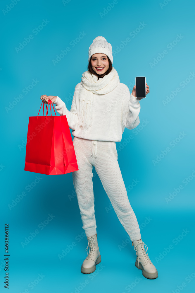 Smiling woman in knitted clothes showing smartphone with blank screen and shopping bags on blue background Smiling woman in knitted clothes showing smartphone with blank screen and shopping bags on blue background