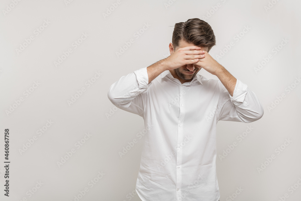 Tired young man in casual white shirt posing isolated on white background, studio portrait. Mock up copy space. Keeping eyes closed, putting both hands on forehead Tired young man in casual white shirt posing isolated on white background, studio portrait. Mock up copy space. Keeping eyes closed, putting both hands on forehead