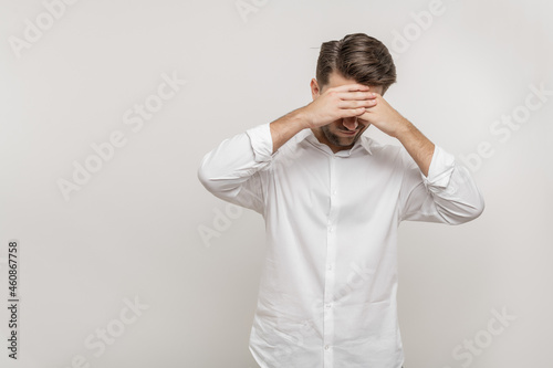 Tired young man in casual white shirt posing isolated on white background, studio portrait. Mock up copy space. Keeping eyes closed, putting both hands on forehead