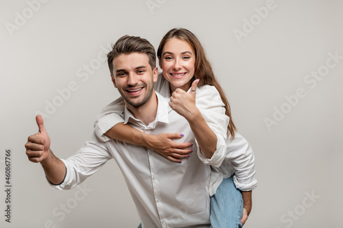 Image of excited couple having fun while man piggybacking joyful woman with thumb up isolated over white background