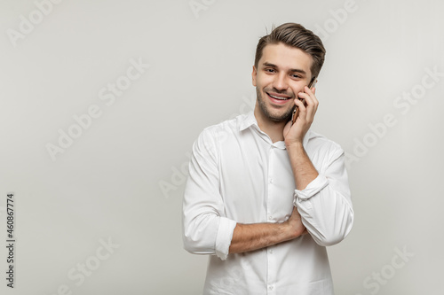 Portrait of a handsome young man in white shirt talking on mobile phone watching at camera isolated over white background