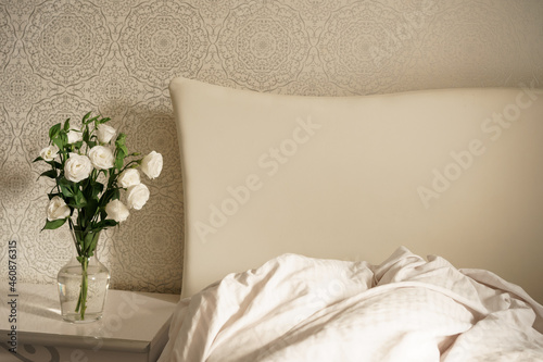 white flowers in a transparent glass vase stand on a bedside table by a bed with a large white headboard. bedroom interior. modern room	
