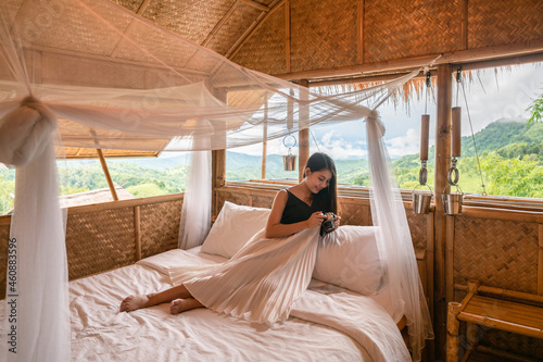 Young asian woman resting and looking at retro camera on the bed in wooden cottage among the mountain