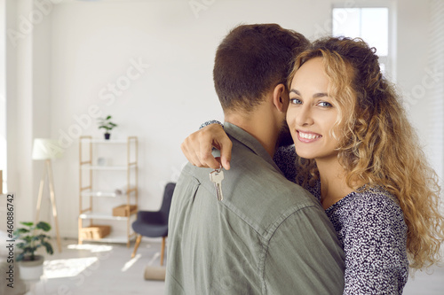 New home new life. Young woman holding keys hugging husband in their new home after buying real estate. Close up of woman in arms of her husband holding key house of her dreams and looking at camera