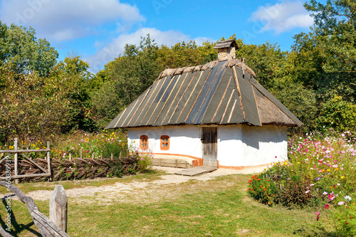 Reconstruction of an old traditional Ukrainian rural house with a thatched roof against the backdrop of a summer garden with a flower bed.