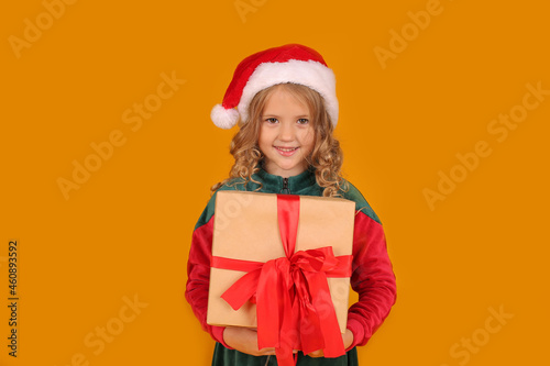 a beautiful blonde girl in a red and green Santa Claus hat dress holds a gift box on a yellow background
