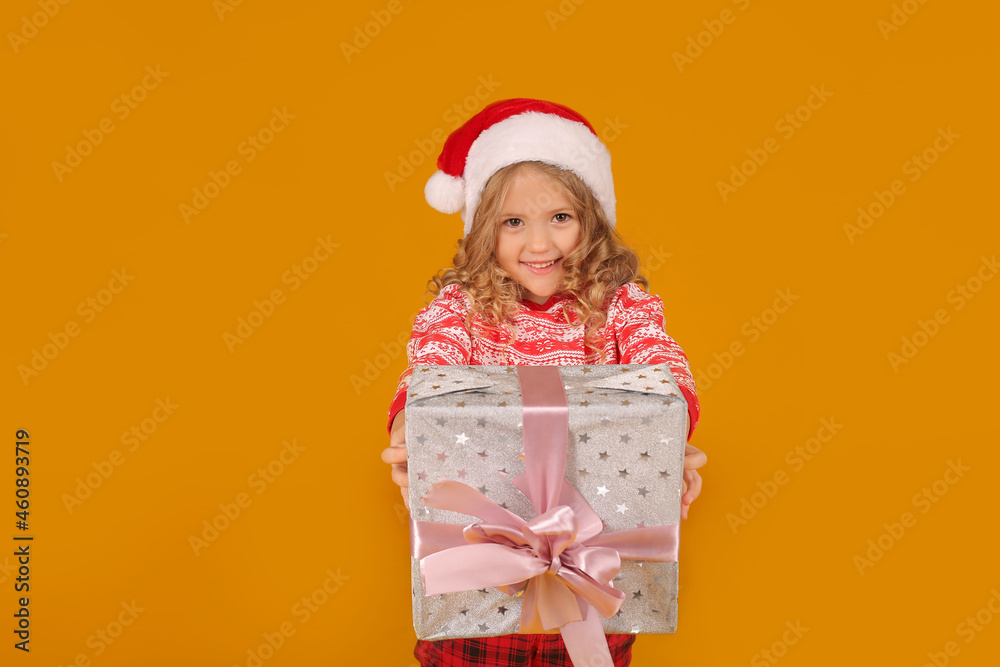 a little beautiful happy blonde girl in a Christmas jumper Santa Claus hat is holding a gift box on a yellow background a little beautiful happy blonde girl in a Christmas jumper Santa Claus hat is holding a gift box on a yellow background