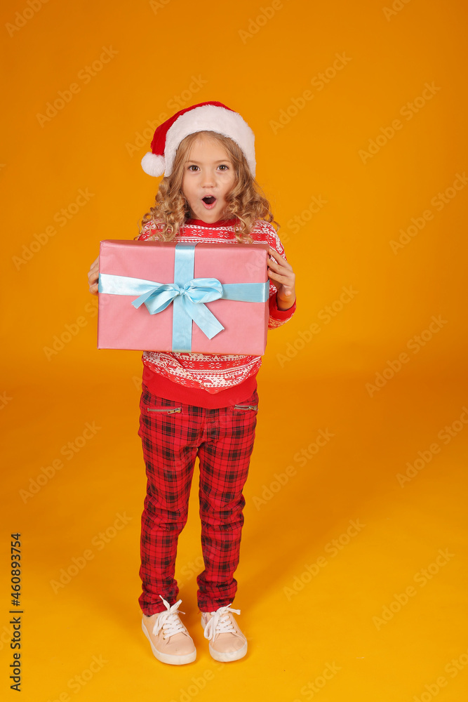 a little beautiful happy blonde girl in a New Year's jumper and plaid trousers with a Santa Claus hat is holding a gift box on a yellow background
