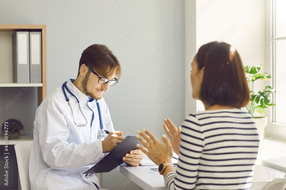 Fototapeta premium Male doctor writes prescription or makes notes in patient's card at medical examination. Doctor listens to woman who talks about health complaints sitting with her back to the camera.