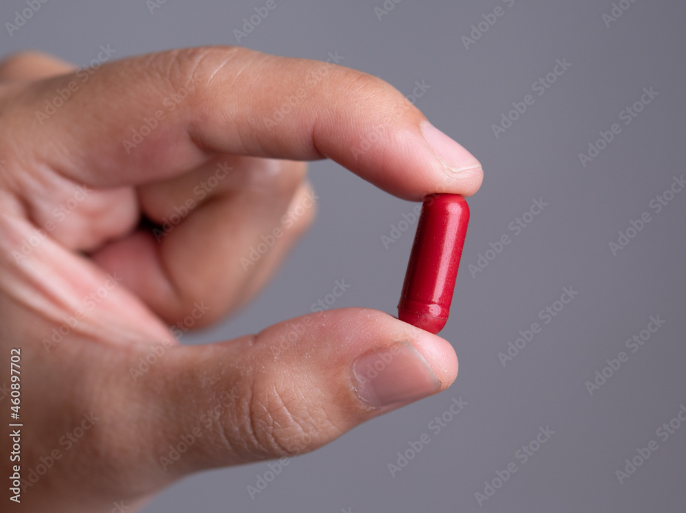 Male hand holding a medical red capsule pill between fingers against gray background Male hand holding a medical red capsule pill between fingers against gray background