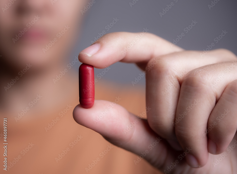 Female hand holding a medical red capsule pill between fingers against gray background Female hand holding a medical red capsule pill between fingers against gray background