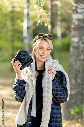 girl with a camera in a city park at work, photographing nature. Girl photographer in a plaid shirt wearing sunglasses in a white sweater