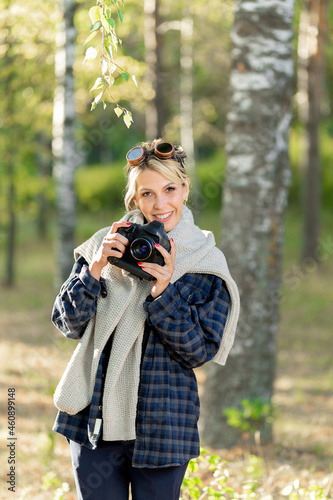 girl with a camera in a city park at work, photographing nature. Girl photographer in a plaid shirt wearing sunglasses in a white sweater