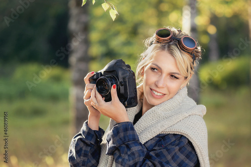 girl with a camera in a city park at work, photographing nature. Girl photographer in a plaid shirt wearing sunglasses in a white sweater