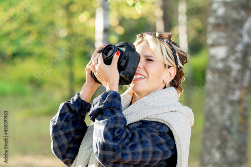 girl with a camera in a city park at work, photographing nature. Girl photographer in a plaid shirt wearing sunglasses in a white sweater