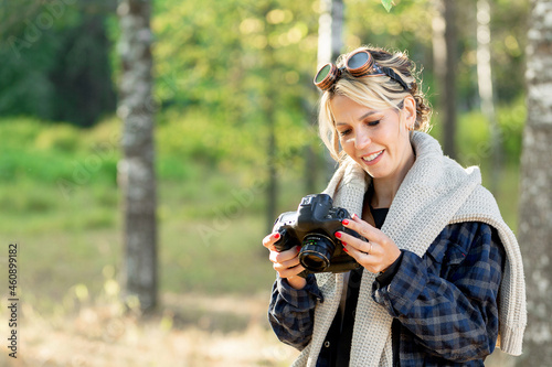girl with a camera in a city park at work, photographing nature. Girl photographer in a plaid shirt wearing sunglasses in a white sweater