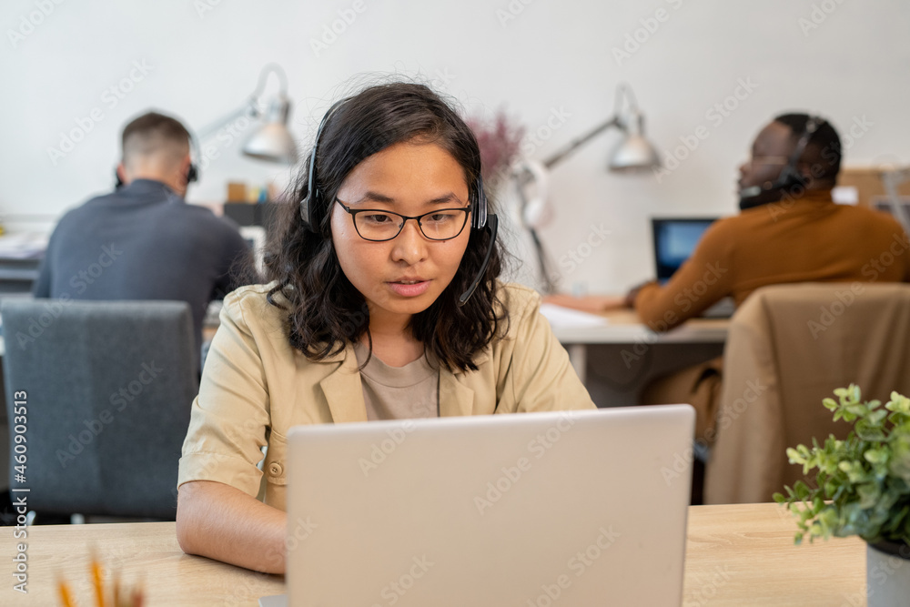 Fototapeta premium Young Asian female in eyeglasses and casualwear consulting clients online in front of laptop in working environment