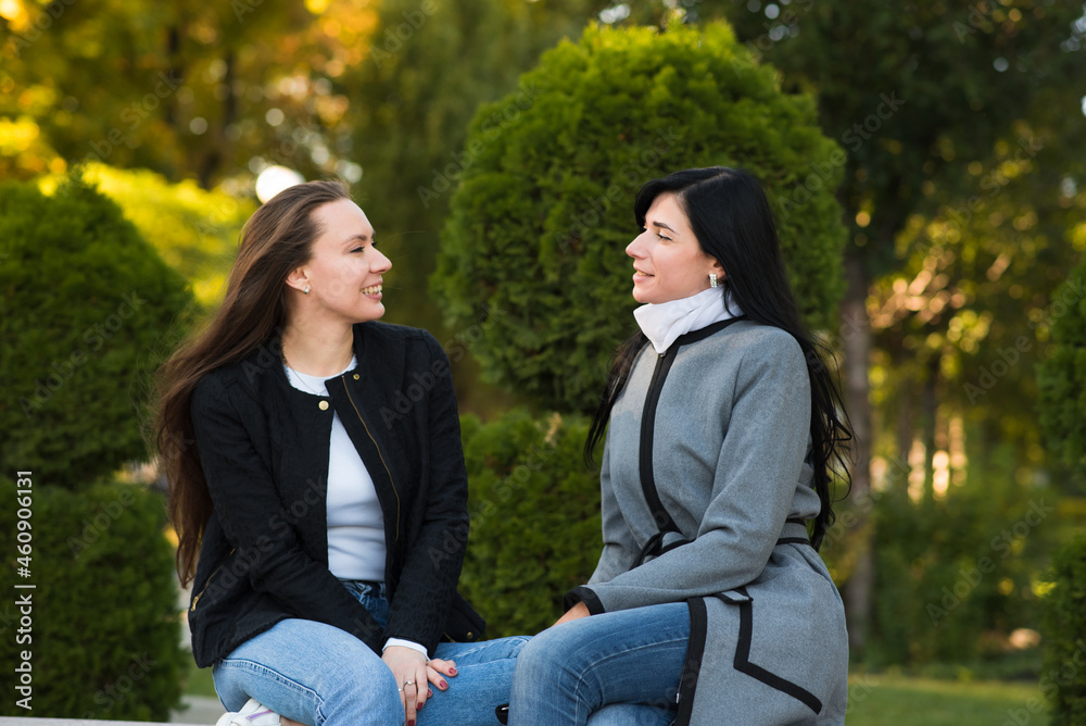 Two Caucasian brunette girls friends enjoying conversation while sitting in sunny autumn park.