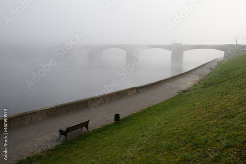 Mysterious perspective of new Volga bridge in thick morning fog in city Tver with benches and green grass on river Volga embankment and nobody else. Horror or silence concept. Russia, Tver.