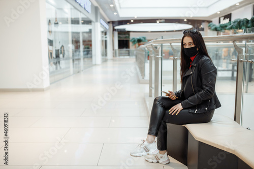 Girl with medical black mask and mobile phone in a shopping center. Coronavirus pandemic. A woman with a mask is standing in a shopping center. A girl in a protective mask is shopping at the mall