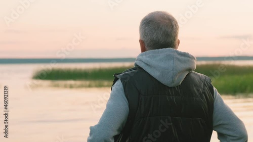 A gray-haired man stands on the shore of a pond and admires nature at sunset on a late autumn evening.