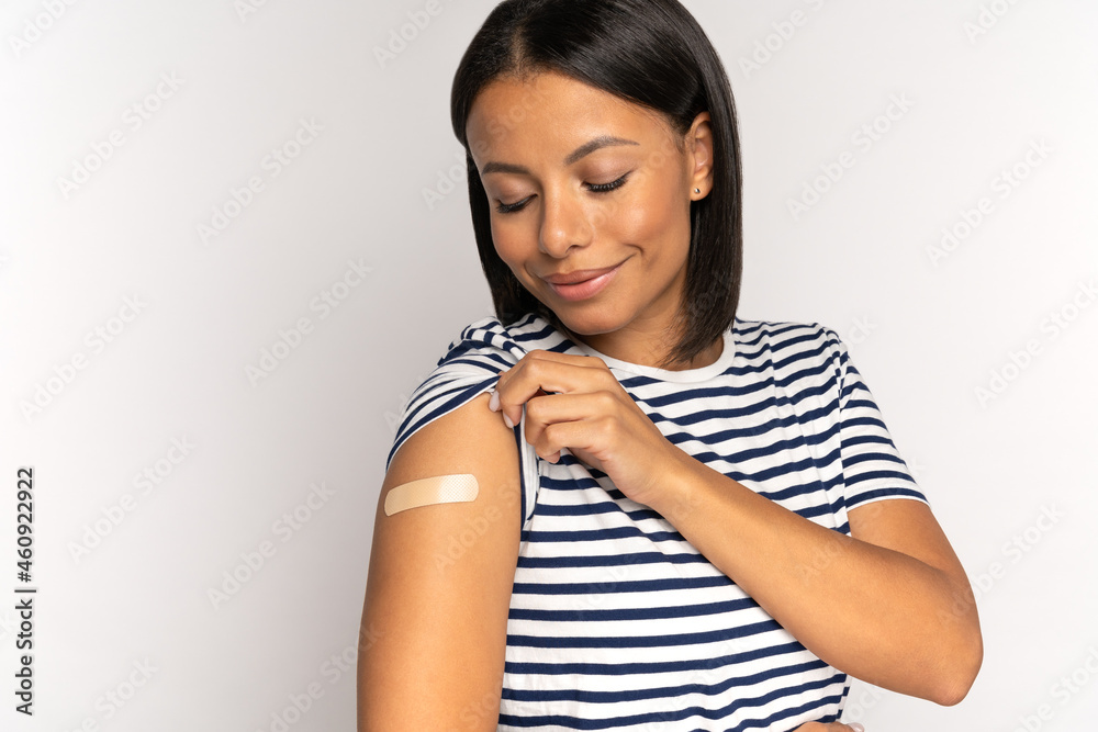 Happy african american woman look at adhesive plaster on shoulder after getting coronavirus vaccine. Young female and covid-10 vaccination injection. Cheerful vaccinated girl. Immunization and health Happy african american woman look at adhesive plaster on shoulder after getting coronavirus vaccine. Young female and covid-10 vaccination injection. Cheerful vaccinated girl. Immunization and health