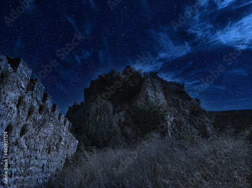 Old ancient castle on the hill at night. Rocky peaks of the ridge in the distant background in full moon light and cloudy sky.