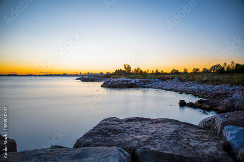 Murais de parede The rocky shoreline and calm, glassy water of Lake Ontario are seen during a colourful sunset in Colonel Samuel Smith Park in Toronto