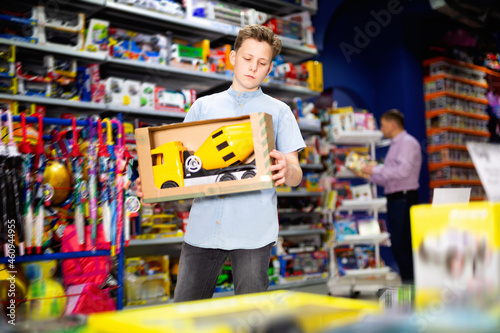 Serious glad cheerful smiling teen boy looking narrowly at yellow plastic car in children toy store