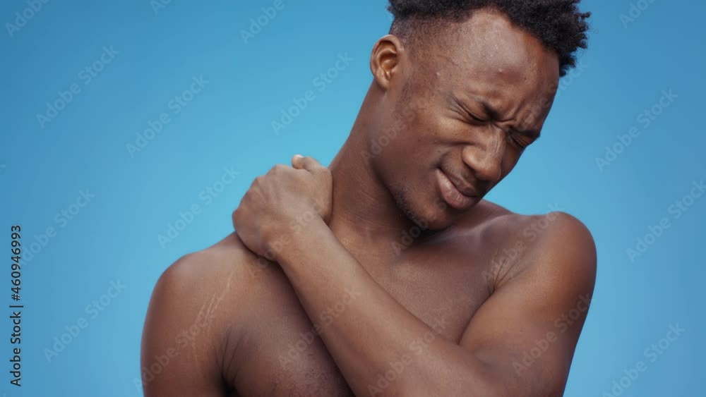 Young upset african american man massaging his painful shoulder, suffering from muscle pain, blue studio background Young upset african american man massaging his painful shoulder, suffering from muscle pain, blue studio background
