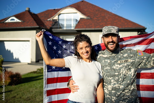 Photography Portrait of husband soldier in uniform on military leave hugging his lovely wife and holding American flag in front of their house