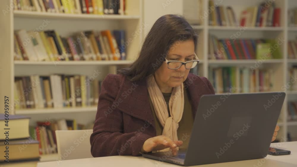 Concentrated old woman in a library using laptop. Close up shot of a serious focused woman sitting on a table and using laptop.