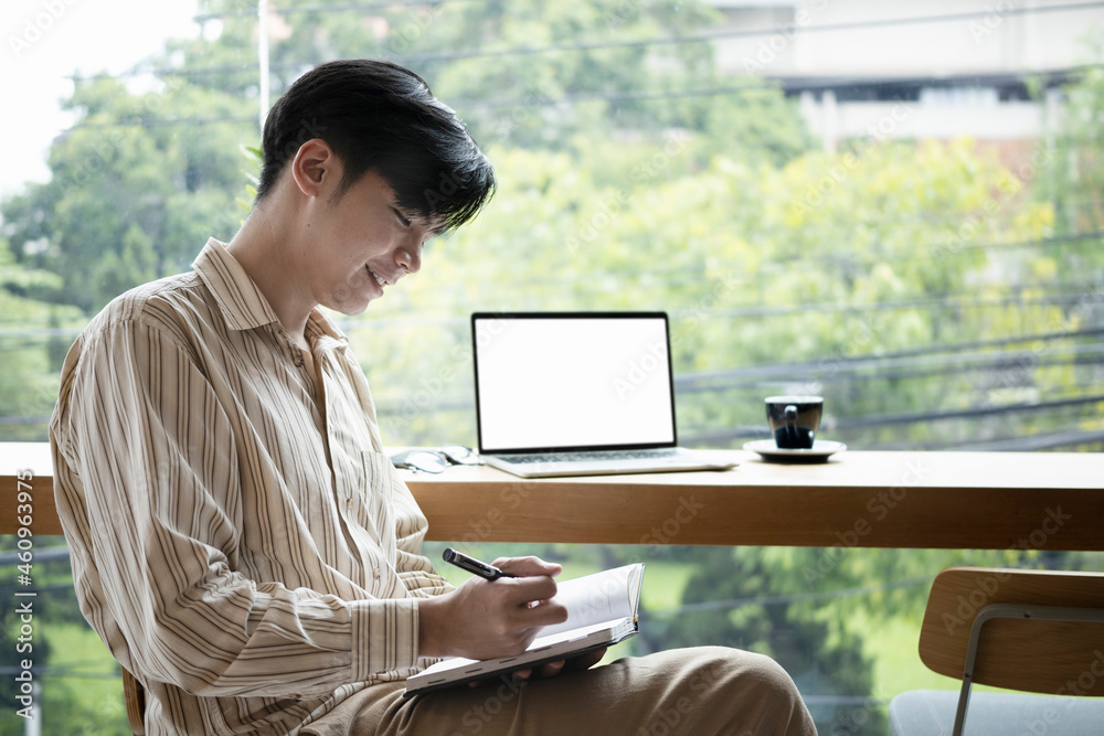 Smiling young Asian man sitting in bright office and writing new idea on notebook. Smiling young Asian man sitting in bright office and writing new idea on notebook.