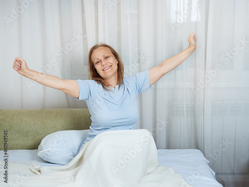 Happy smiling senior woman in blue pajamas stretching in the morning after sleeping in her bed.