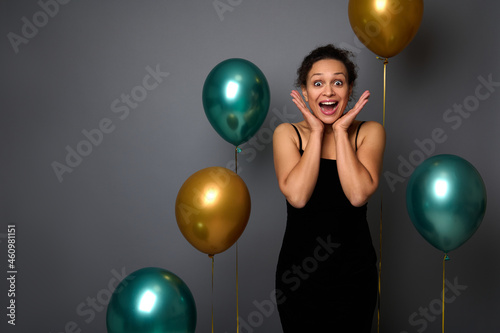 Beautiful woman in black dress holds her hands on her cheeks, poses with opened mouth expressing happiness, surprise. Birthday, Christmas and events concepts on gray background with copy space for ad