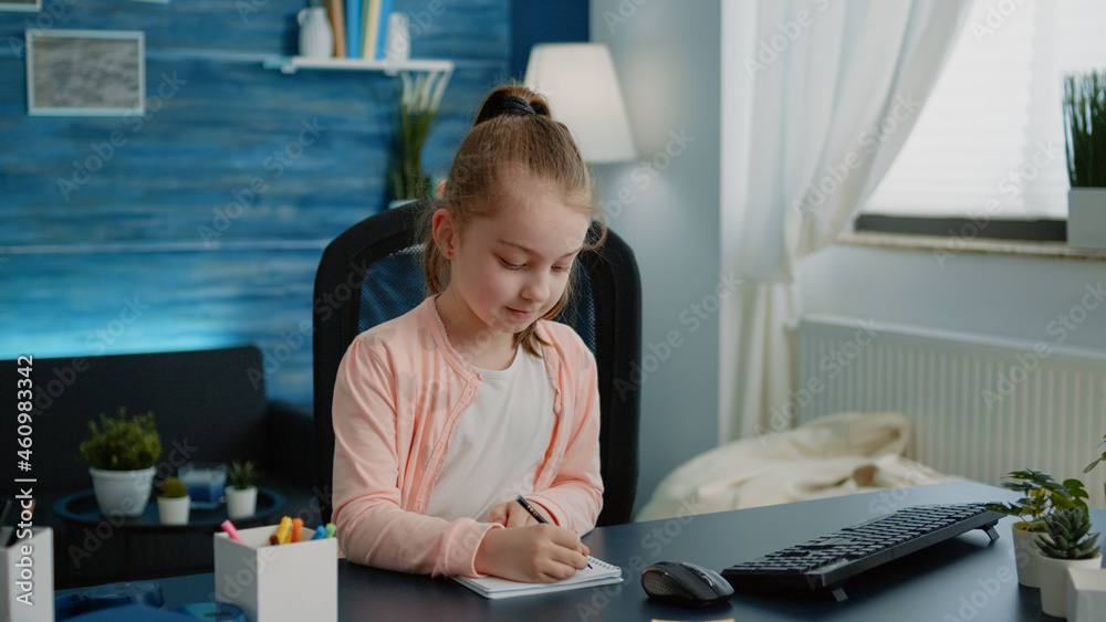 Schoolgirl writing on notebook with pen for homework. Little child concentrated on task for online remote lessons while sitting at desk with computer and textbook. Homeschooled pupil Schoolgirl writing on notebook with pen for homework. Little child concentrated on task for online remote lessons while sitting at desk with computer and textbook. Homeschooled pupil