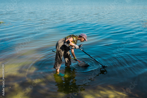  A woman in camouflage clothes collects metal in the river with a metal detector on a sunny day. High quality photo