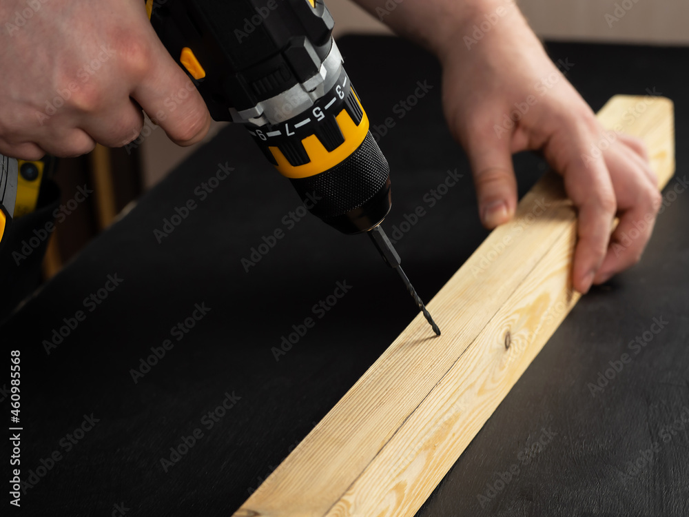 A carpenter drills a hole in a wooden board with an electric drill. Close-up of hands of a foreman at work. Working environment in a carpentry workshop. Cordless cordless drill - screwdriver with dril