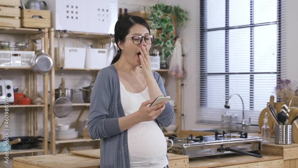 sleepy pregnant woman is yawning while standing in the kitchen at home and phubbing on her smartphone in the morning.