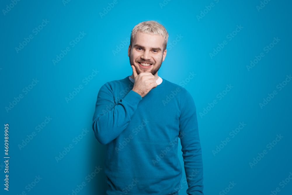 Man with blonde hair on blue wall is touching chin. Smiling man. Front view. Handsome man. Positive person.