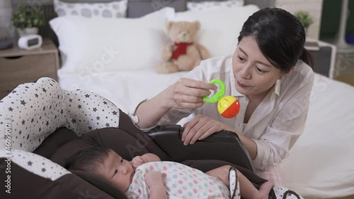 asian newborn infant is getting upset in the bouncer while its mommy is trying to playing with her by shaking a handheld rattle in the bedroom at home. photo