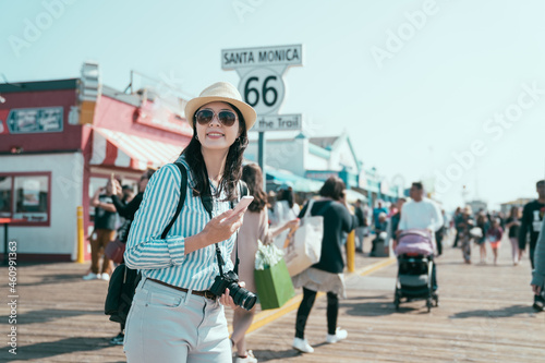 end of route 66 on santa monica pier california. happy smiling young asian chinese woman backpacker holding smartphone and camera joyful looking aside under sunshine. pretty tourist enjoy sun flare photo